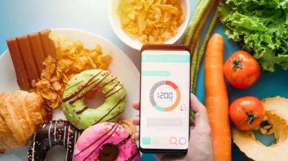 A hand holds a smartphone displaying calorie data next to colorful donuts, pastries, and fresh vegetables.